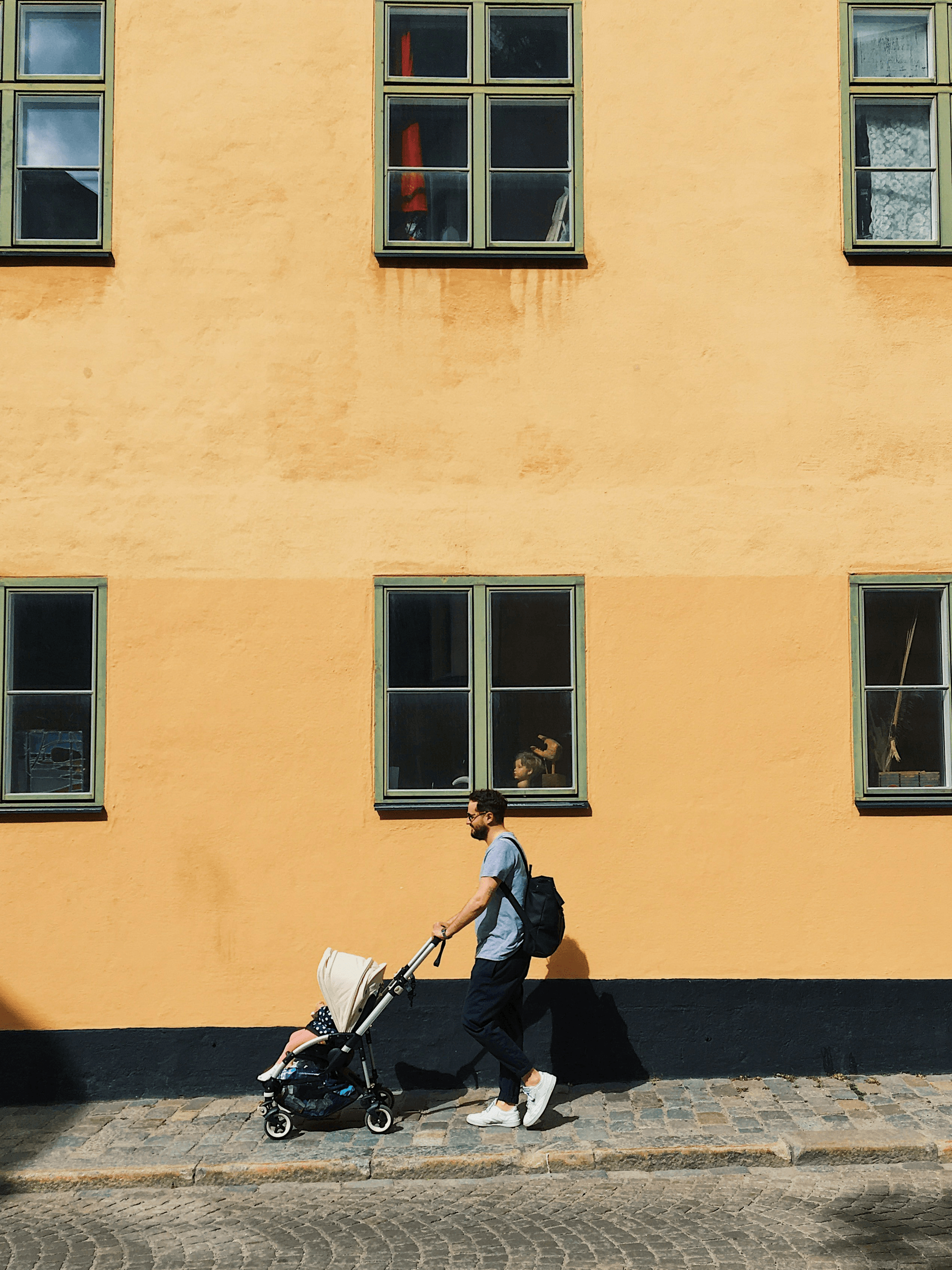 Parent pushing a stroller past an ochre wall and green windows — Stockholm summer ease.